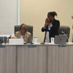 Four women in a meeting room applaud; one seated with a bouquet stands beside laptops, others stand and clap at the long conference table.