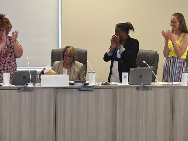 Four women in a meeting room applaud; one seated with a bouquet stands beside laptops, others stand and clap at the long conference table.