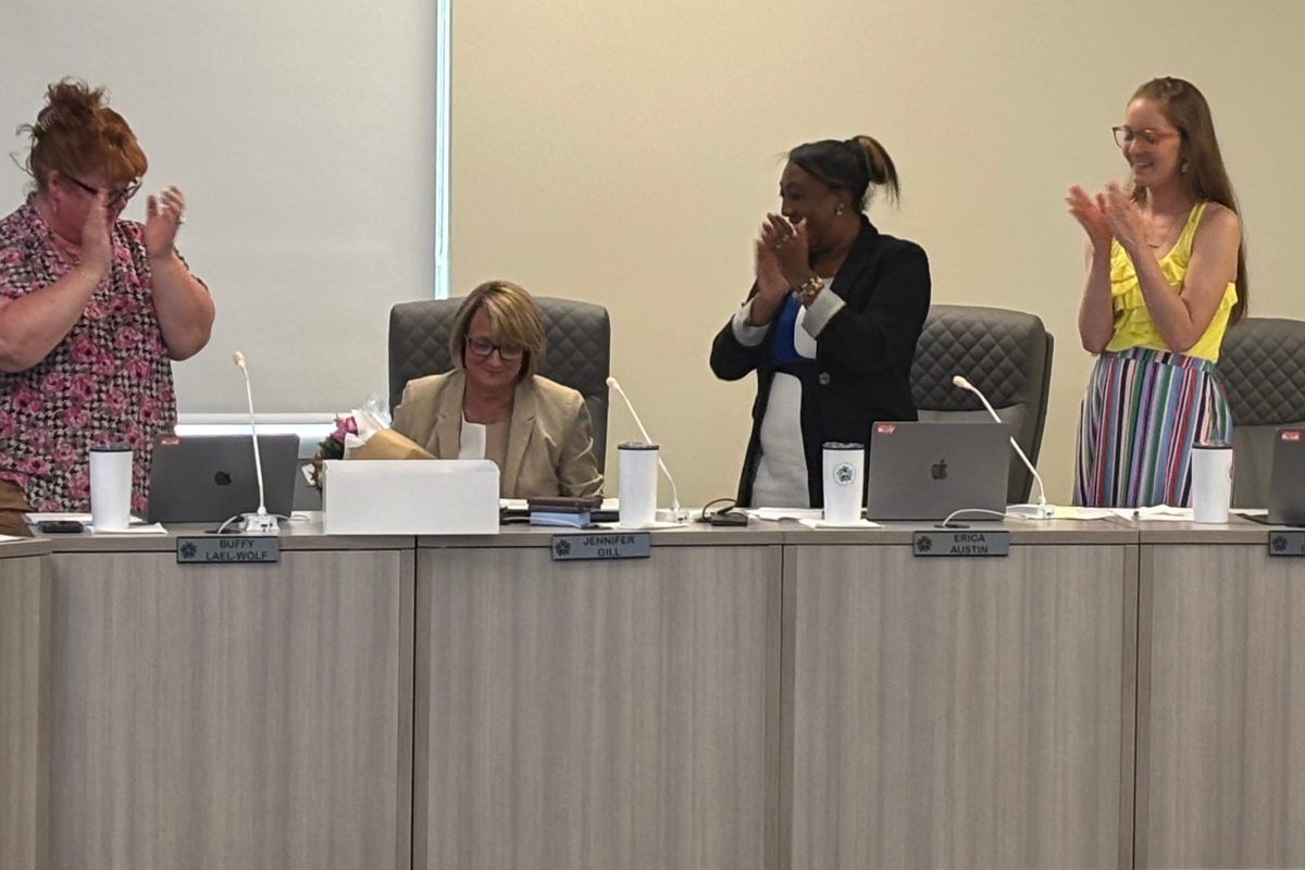 Four women in a meeting room applaud; one seated with a bouquet stands beside laptops, others stand and clap at the long conference table.