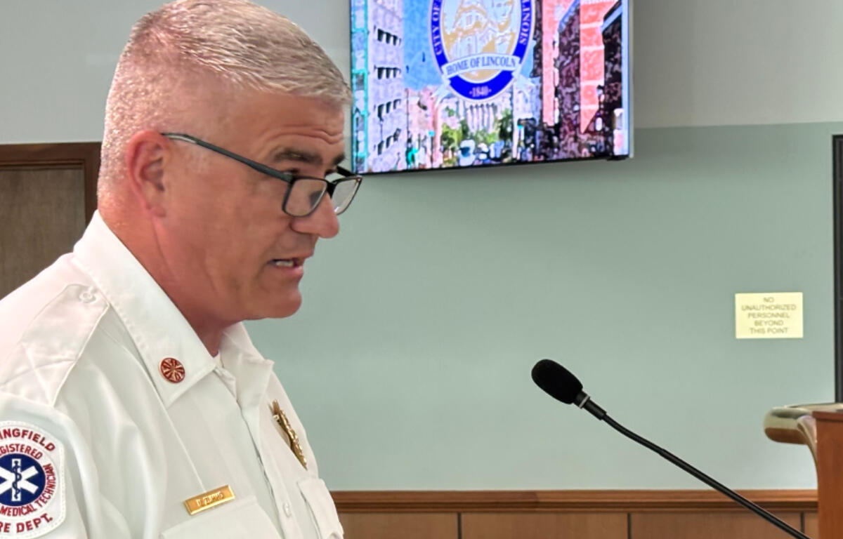 Male emergency services officer speaking at a podium with a microphone, wearing a white uniform and badges in a meeting room.