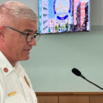 Male emergency services officer speaking at a podium with a microphone, wearing a white uniform and badges in a meeting room.