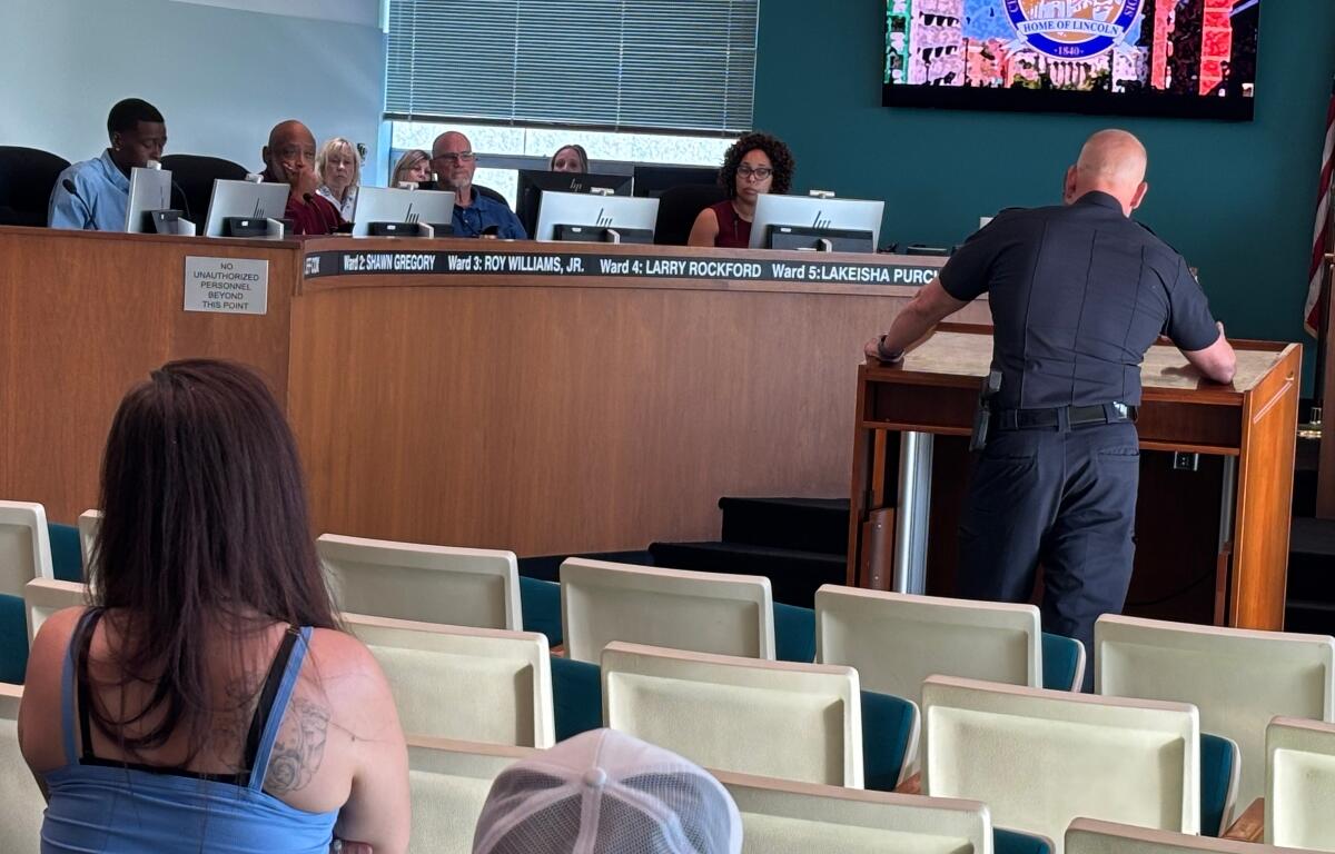 Uniformed officer at a wooden lectern addressing a city council meeting, with council members behind a long desk and audience seating in the foreground.