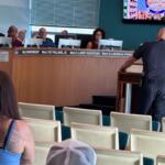 Uniformed officer at a wooden lectern addressing a city council meeting, with council members behind a long desk and audience seating in the foreground.
