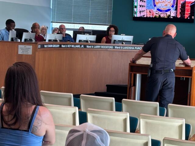 Uniformed officer at a wooden lectern addressing a city council meeting, with council members behind a long desk and audience seating in the foreground.