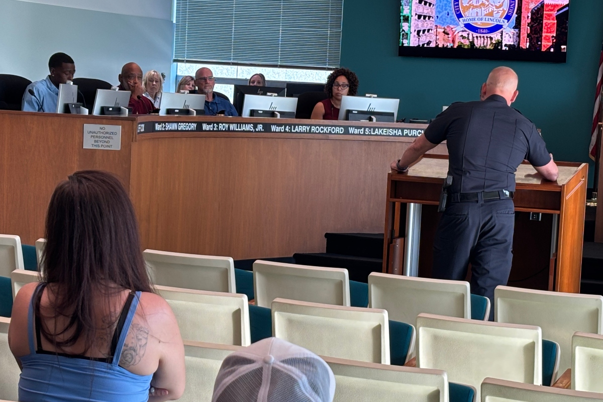 Uniformed officer at a wooden lectern addressing a city council meeting, with council members behind a long desk and audience seating in the foreground.