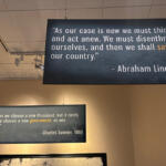 Two hanging blue quote boards on a museum wall; Lincoln (1862) quote about thinking anew and saving the country, and Sumner (1860) quote about government.
