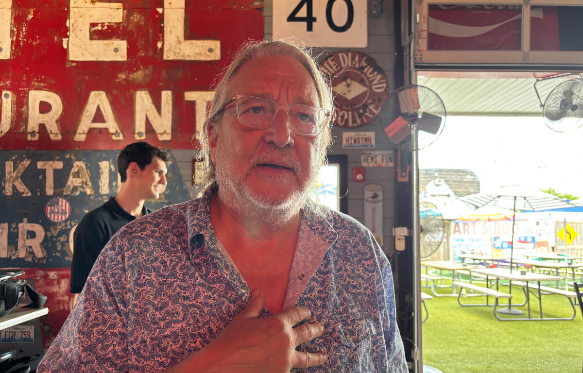 Older man with glasses and a white beard stands inside a rustic eatery, hand on chest, with a weathered red sign behind him and outdoor picnic tables visible outside.