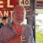 Older man with glasses and a white beard stands inside a rustic eatery, hand on chest, with a weathered red sign behind him and outdoor picnic tables visible outside.