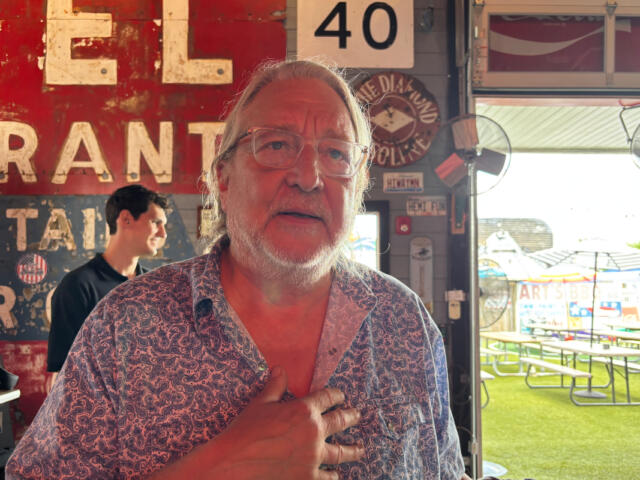 Older man with glasses and a white beard stands inside a rustic eatery, hand on chest, with a weathered red sign behind him and outdoor picnic tables visible outside.