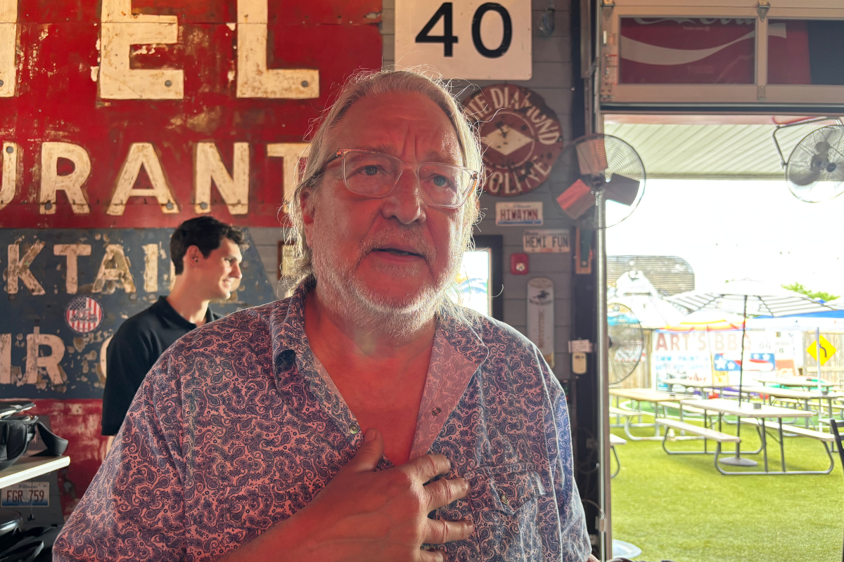 Older man with glasses and a white beard stands inside a rustic eatery, hand on chest, with a weathered red sign behind him and outdoor picnic tables visible outside.