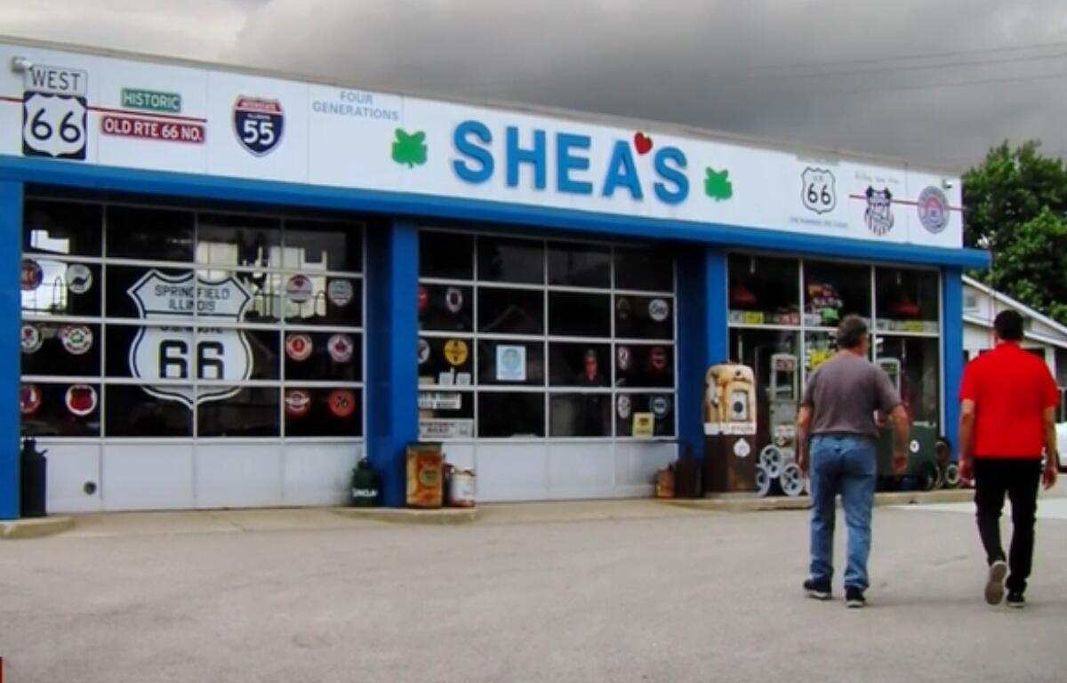 Front of Shea's shop along Route 66, blue trim, large windows with decals; two men approach the entrance.