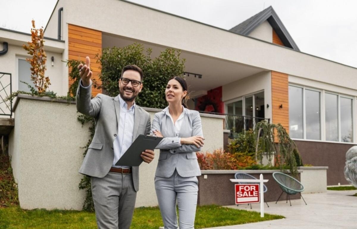 Two real estate agents in gray suits stand outside a modern house with a 'For Sale' sign, discussing the property.