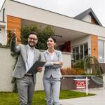 Two real estate agents in gray suits stand outside a modern house with a 'For Sale' sign, discussing the property.
