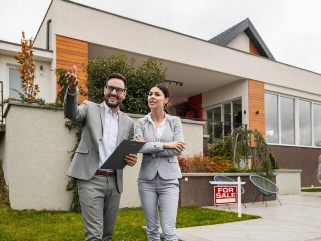 Two real estate agents in gray suits stand outside a modern house with a 'For Sale' sign, discussing the property.