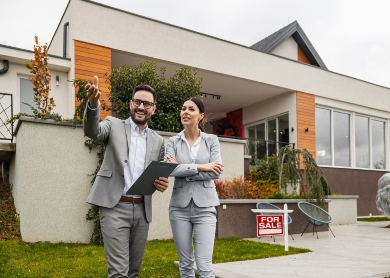 Two real estate agents in gray suits stand outside a modern house with a 'For Sale' sign, discussing the property.
