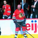 Center ice hockey player in a red Chicago Blackhawks jersey (number 10) smiles while holding a stick near the boards.