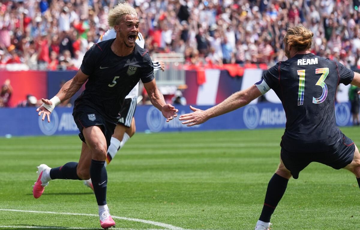 USA national team players celebrate a goal on the green pitch as teammates reach out to hug, crowd cheering in the stands behind.