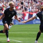 USA national team players celebrate a goal on the green pitch as teammates reach out to hug, crowd cheering in the stands behind.