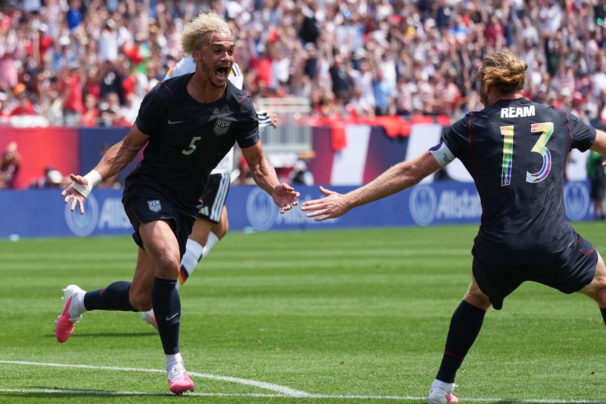USA national team players celebrate a goal on the green pitch as teammates reach out to hug, crowd cheering in the stands behind.