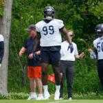 Three American football players in white jerseys (numbers 93, 99, and 50) stand on a grassy practice field with coaches nearby.