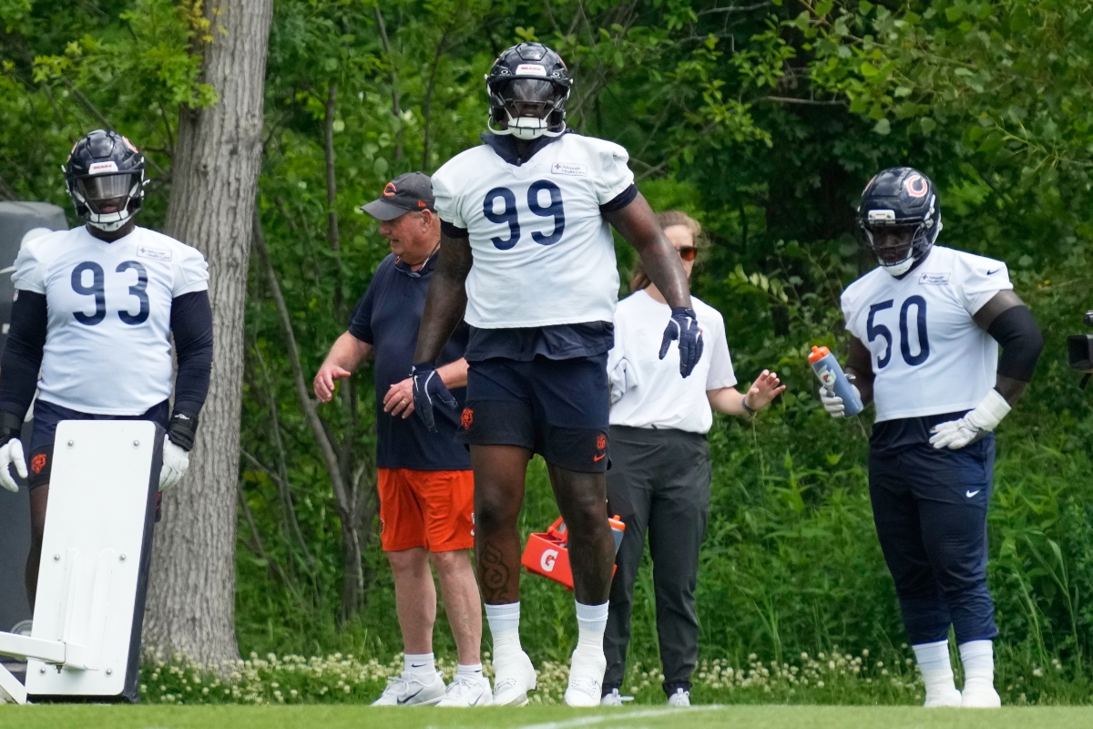 Three American football players in white jerseys (numbers 93, 99, and 50) stand on a grassy practice field with coaches nearby.