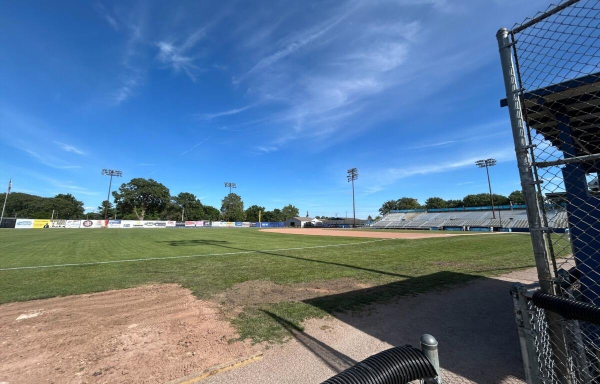 Baseball field with infield dirt, green grass, and empty stands under a clear blue sky, viewed from the dugout area behind a chain-link fence.