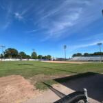 Baseball field with infield dirt, green grass, and empty stands under a clear blue sky, viewed from the dugout area behind a chain-link fence.