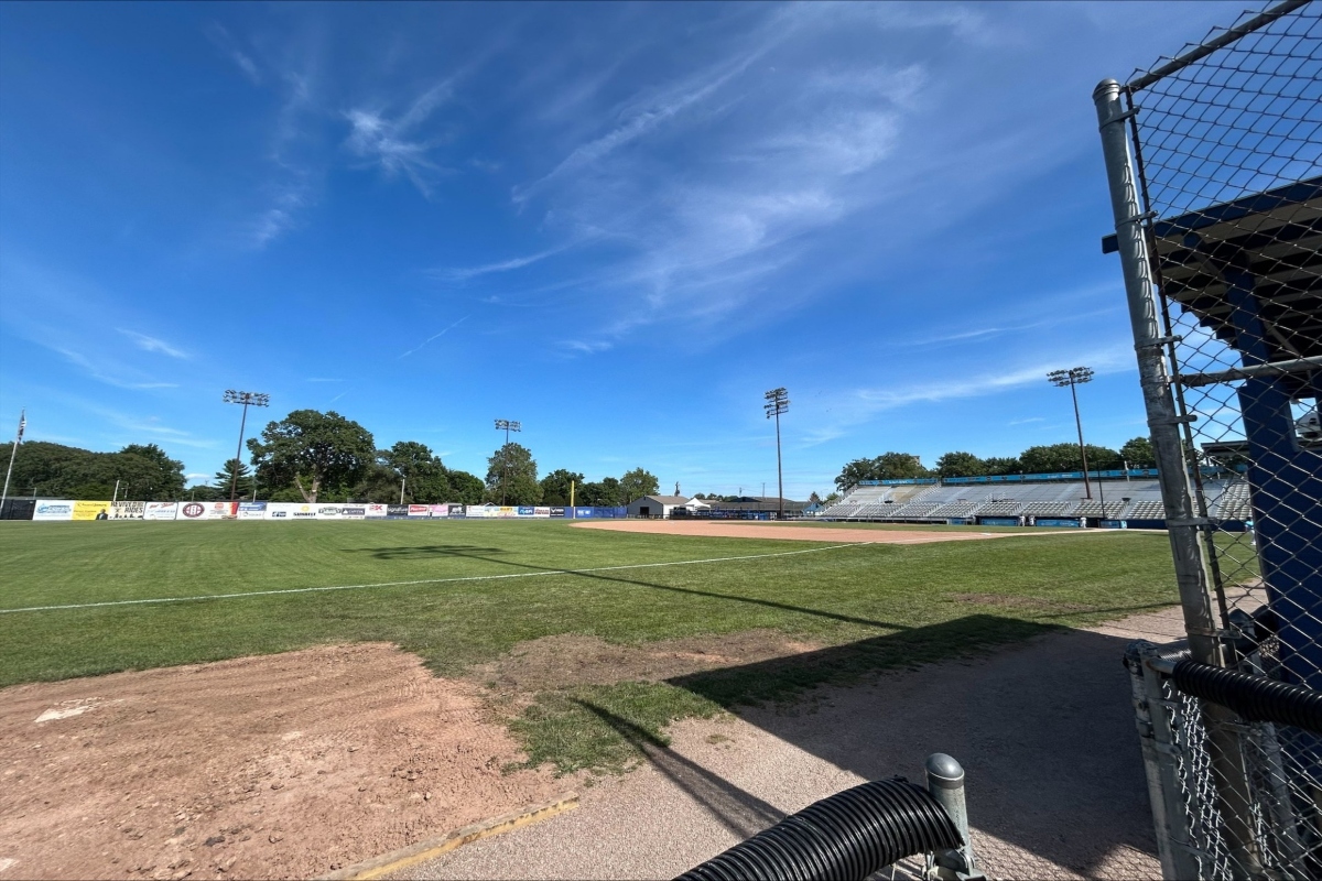 Baseball field with infield dirt, green grass, and empty stands under a clear blue sky, viewed from the dugout area behind a chain-link fence.