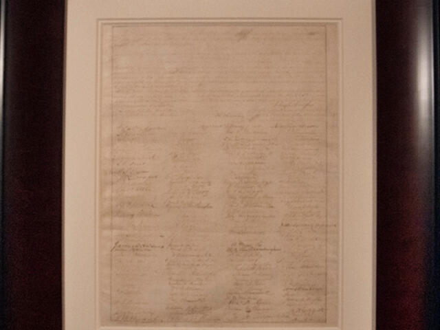 Framed parchment with dense handwritten text displayed on a wall/gallery setting.