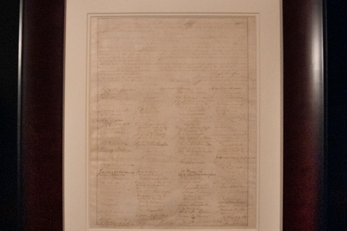 Framed parchment with dense handwritten text displayed on a wall/gallery setting.