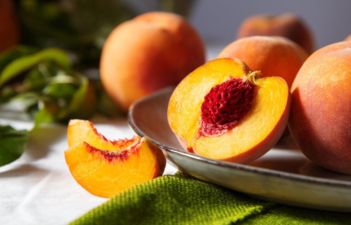 Halved peach with bright orange flesh and red pit on a metal platter, surrounded by whole peaches in a still life setting.