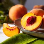 Halved peach with bright orange flesh and red pit on a metal platter, surrounded by whole peaches in a still life setting.