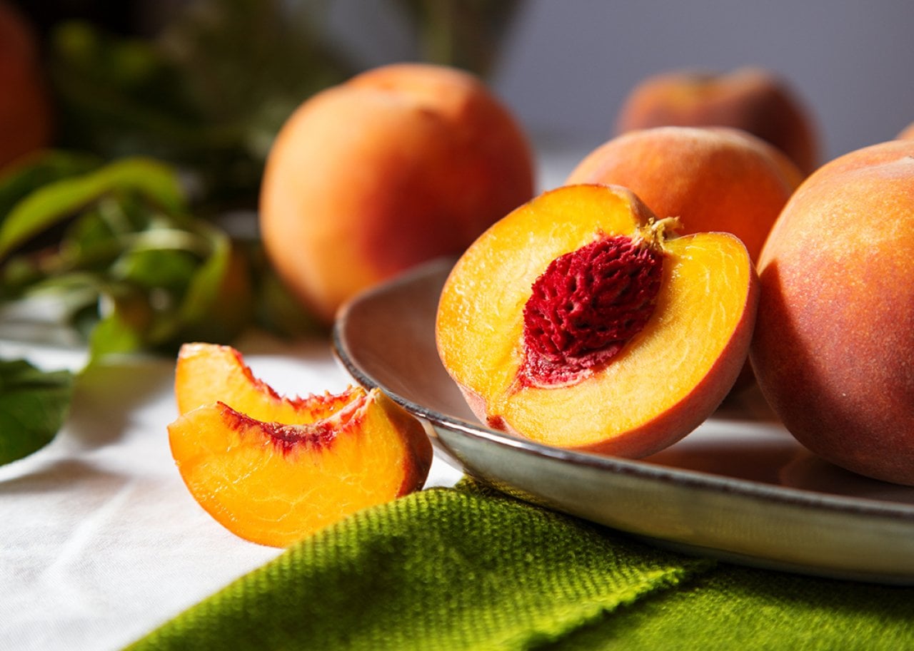 Halved peach with bright orange flesh and red pit on a metal platter, surrounded by whole peaches in a still life setting.