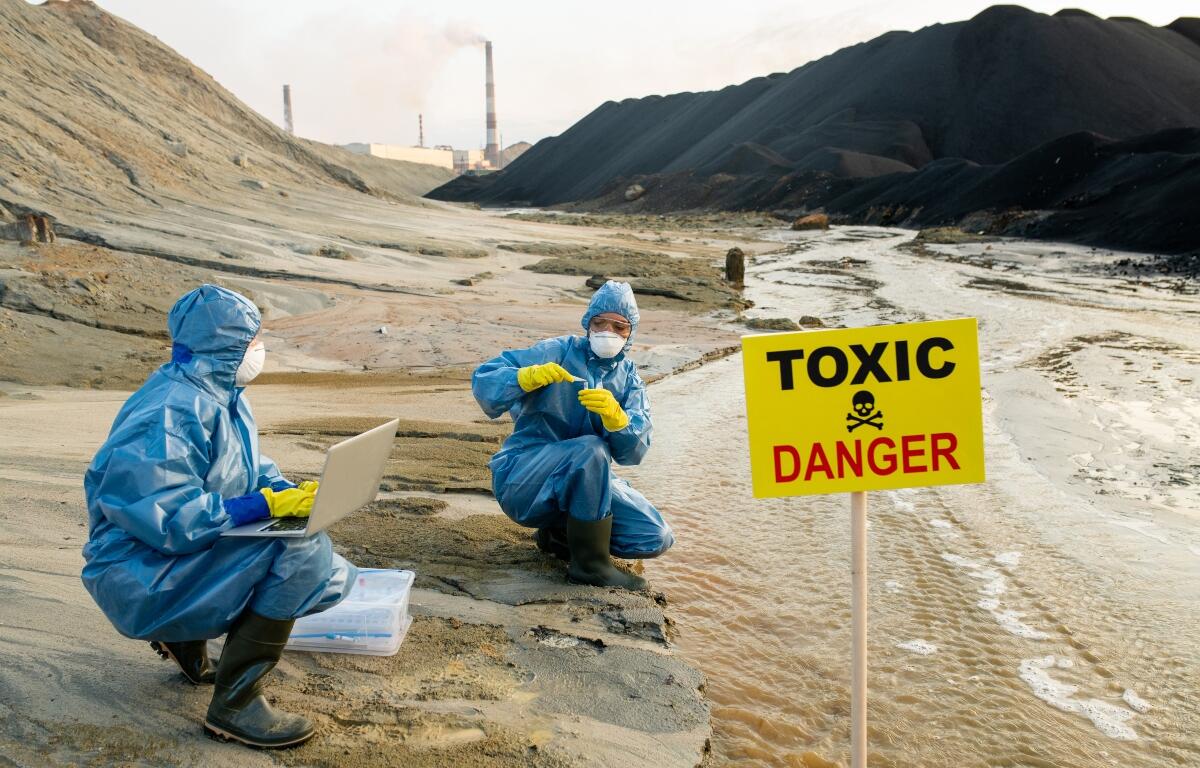 Two young female ecologists in protective coveralls, eyeglasses and respirators studying characteristics of toxic water on riverbank