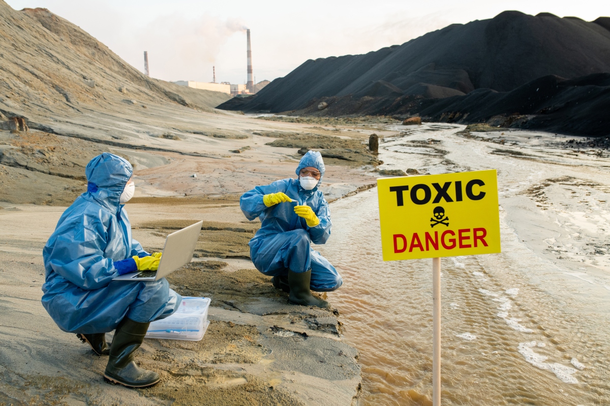 Two young female ecologists in protective coveralls, eyeglasses and respirators studying characteristics of toxic water on riverbank