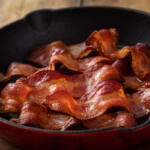 Crispy bacon slices arranged in a red cast-iron skillet on a wooden table.