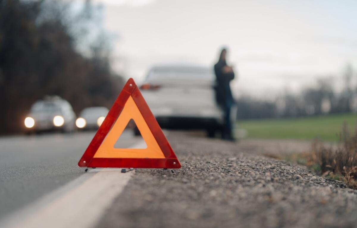 Orange warning triangle on the road shoulder with a blurred white car and person in the background.