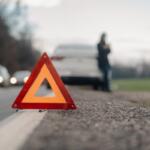 Orange warning triangle on the road shoulder with a blurred white car and person in the background.
