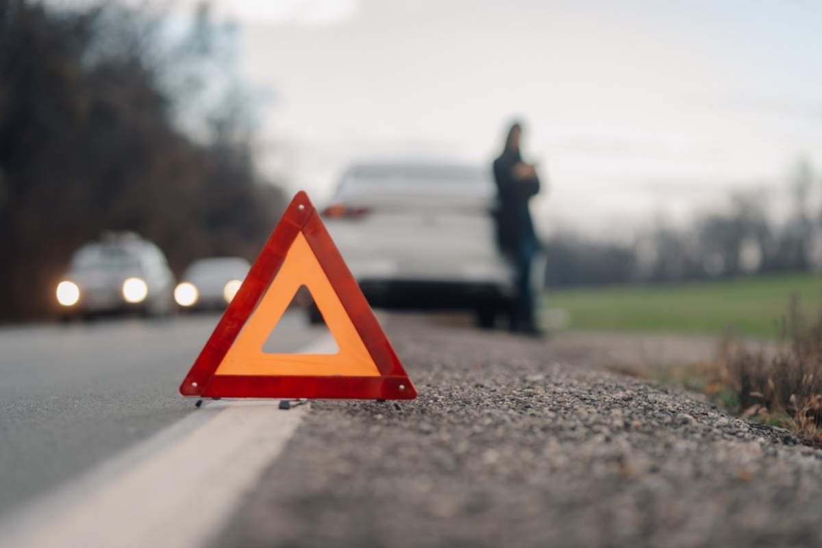 Orange warning triangle on the road shoulder with a blurred white car and person in the background.