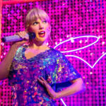 Singer performing on a colorful neon-lit stage in a sparkling blue sequined dress, holding a glittery microphone.