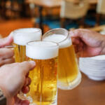 Friends raising beer mugs in a toast at a bar or restaurant, foam on top and warm lighting in the background.