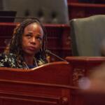 Woman with braided hair seated behind a wood desk, speaking into a microphone in a formal legislative chamber.