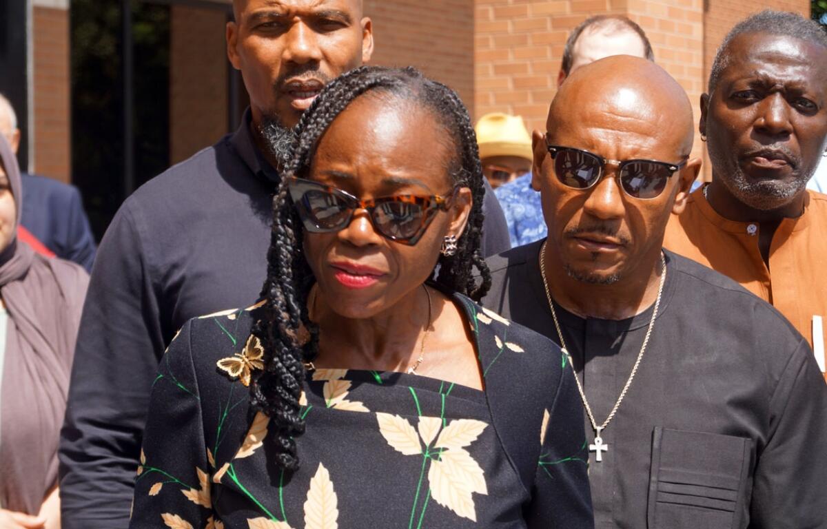 A woman in a black floral dress wearing sunglasses leads a small group outdoors, with men nearby wearing dark clothing and a brick building in the background.