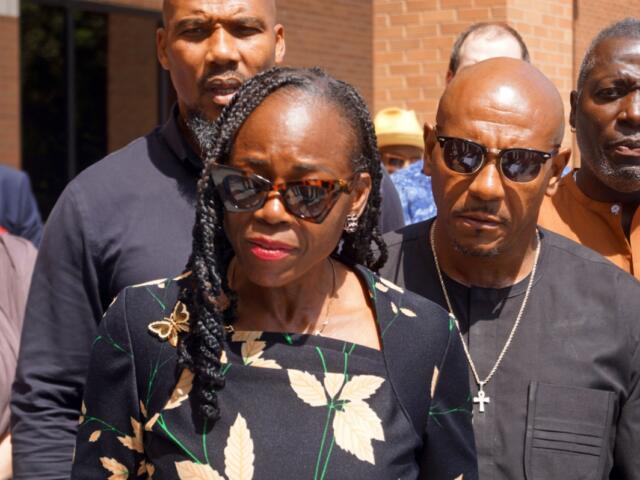 A woman in a black floral dress wearing sunglasses leads a small group outdoors, with men nearby wearing dark clothing and a brick building in the background.