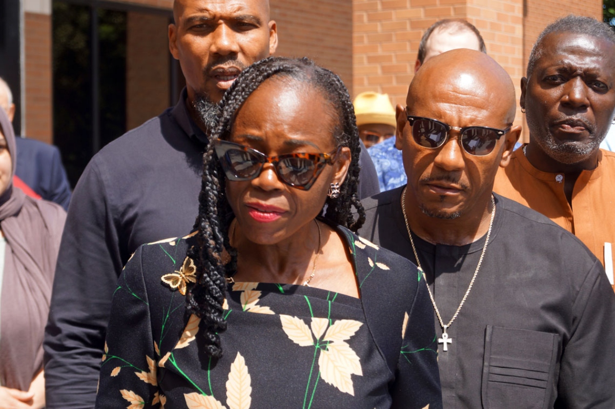 A woman in a black floral dress wearing sunglasses leads a small group outdoors, with men nearby wearing dark clothing and a brick building in the background.