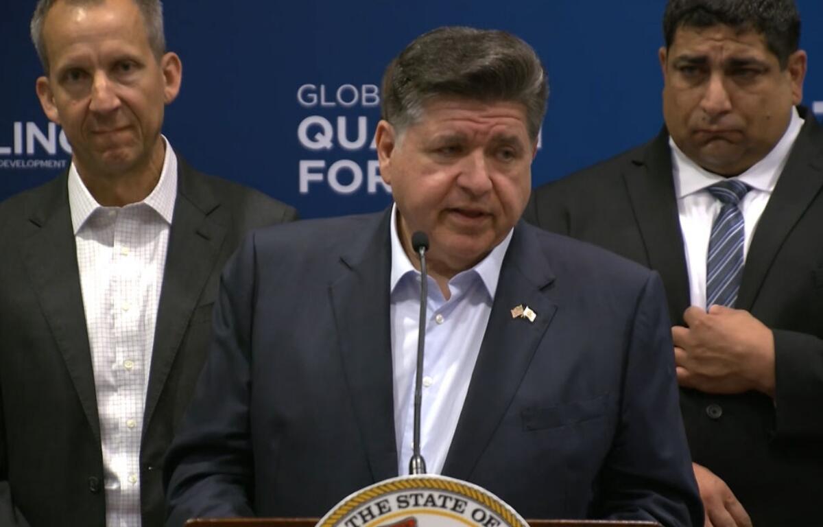 Man at a podium giving a formal speech with a state seal on the lectern and two men in suits behind him.