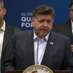 Man at a podium giving a formal speech with a state seal on the lectern and two men in suits behind him.