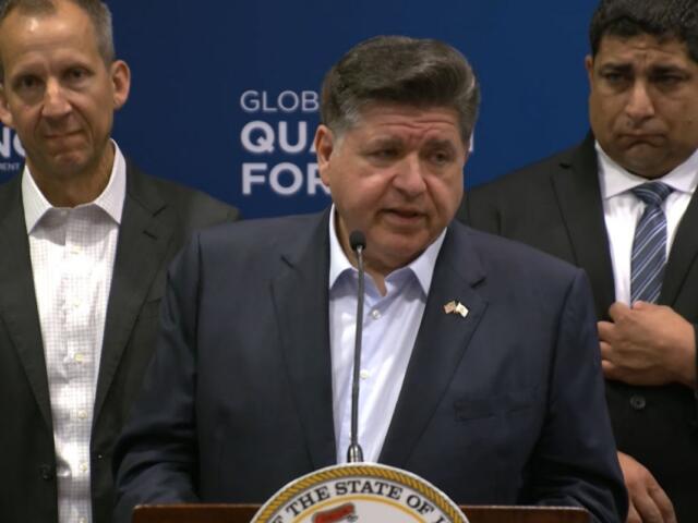 Man at a podium giving a formal speech with a state seal on the lectern and two men in suits behind him.