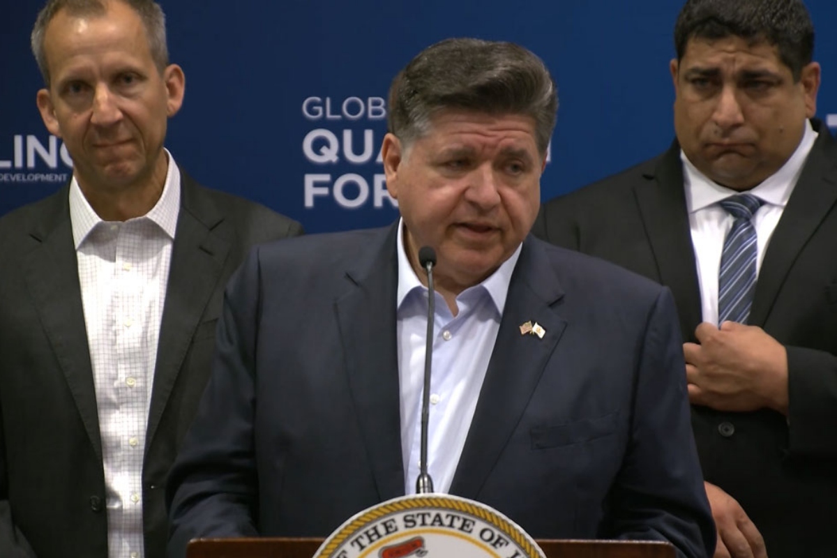 Man at a podium giving a formal speech with a state seal on the lectern and two men in suits behind him.