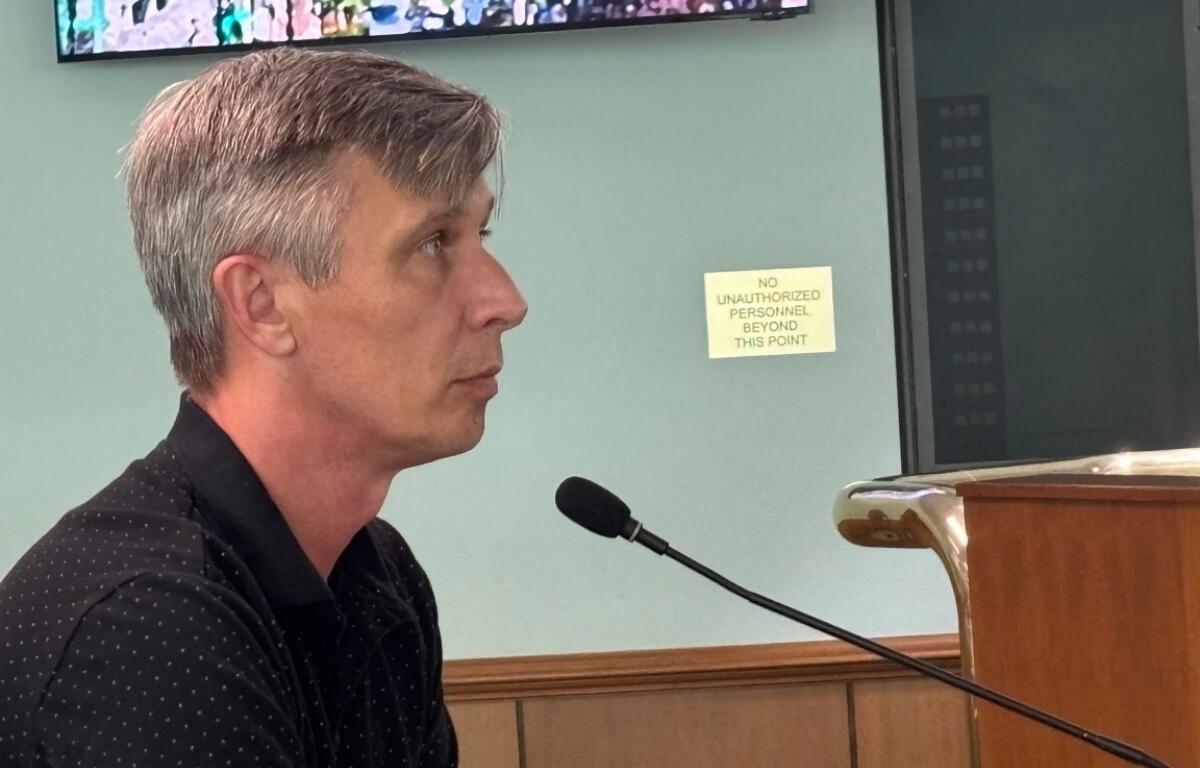 Side profile of a man with gray hair speaking into a microphone at a wooden lectern; a sign behind him reads 'No unauthorized personnel beyond this point'